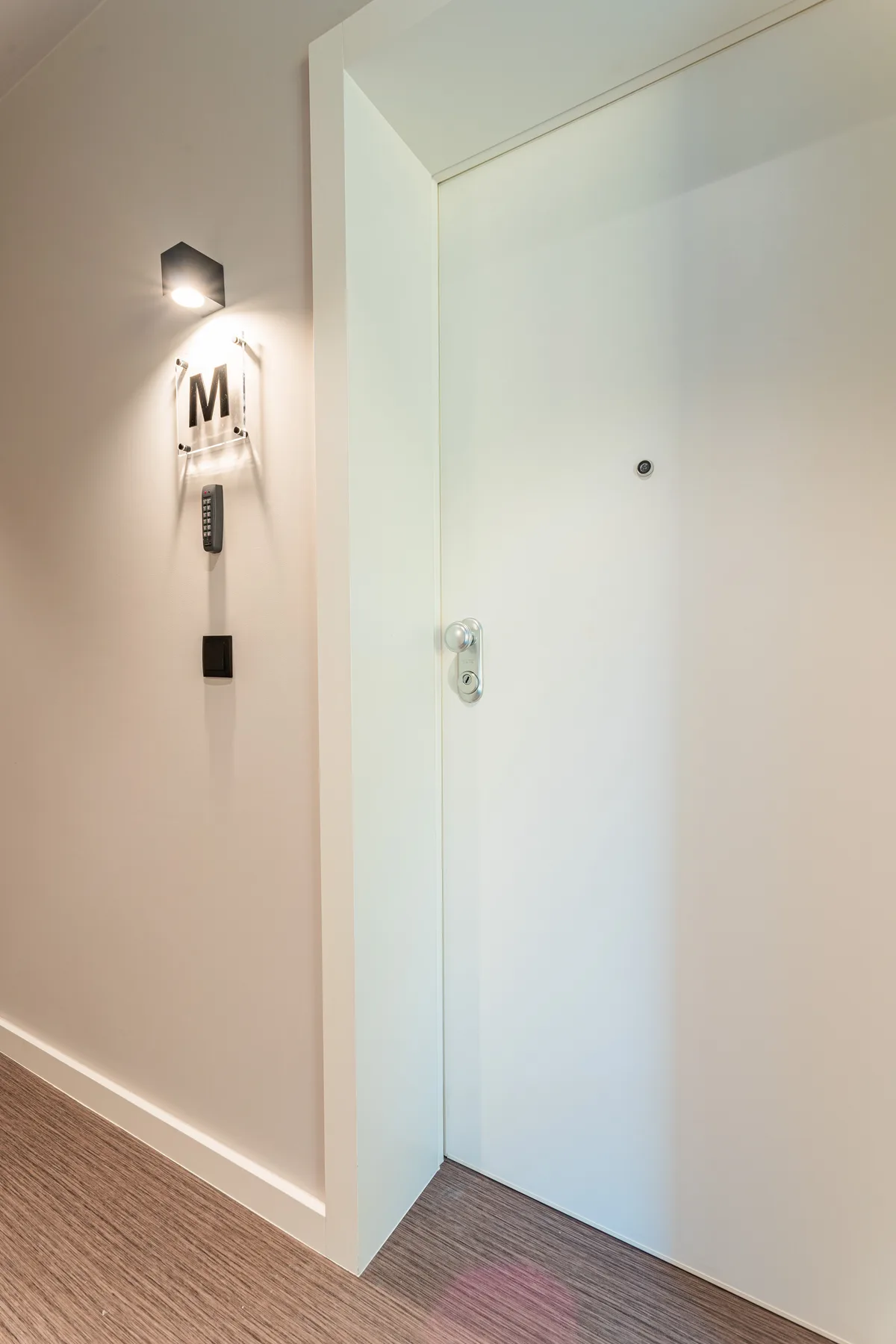 A white entry door opens onto a hallway with a wall light and controls.