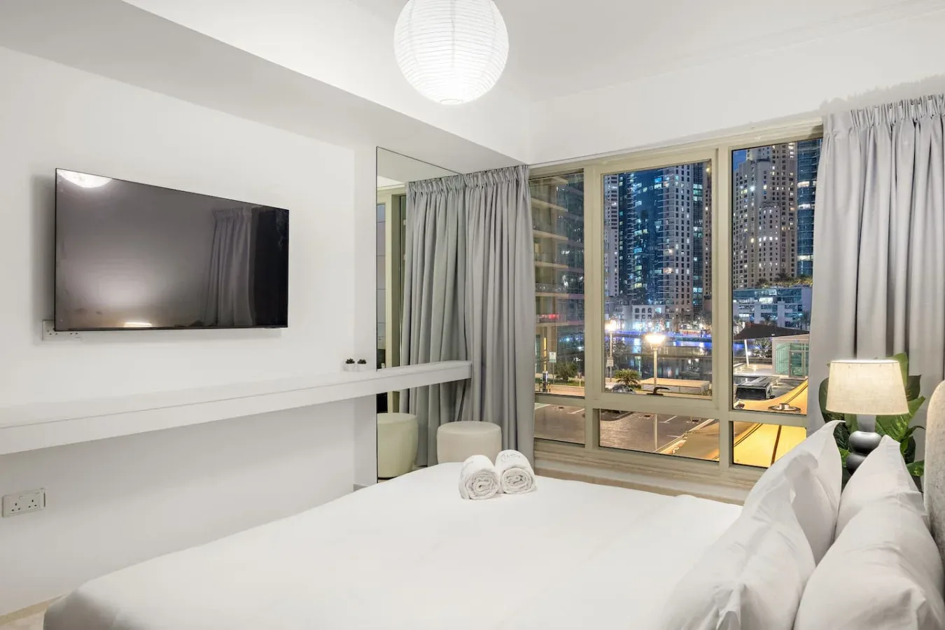 Un dormitorio luminoso con un televisor en la pared y ventanas amplias con vistas a las luces de la ciudad.