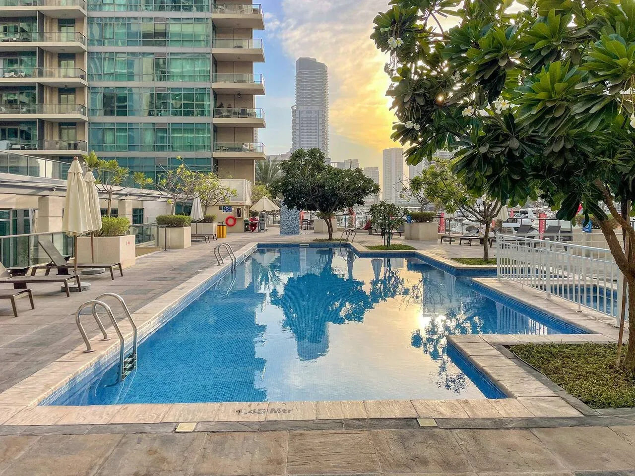 Una piscina al aire libre está junto a un edificio moderno con fachada de vidrio y una zona ajardinada con asientos.