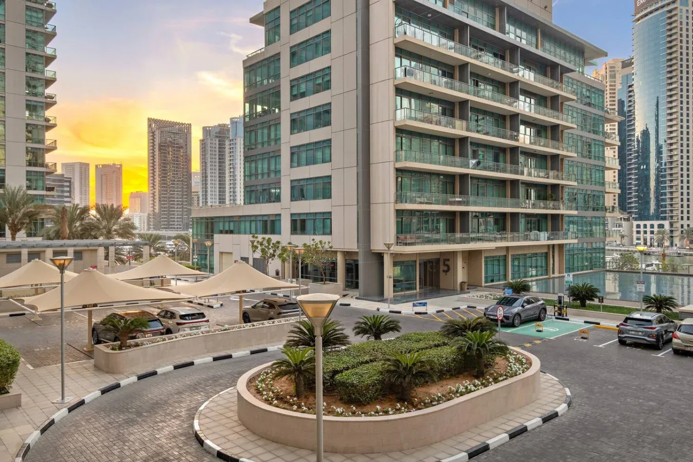 Edificios modernos de gran altura rodean un patio ajardinado y una entrada pavimentada al atardecer.
