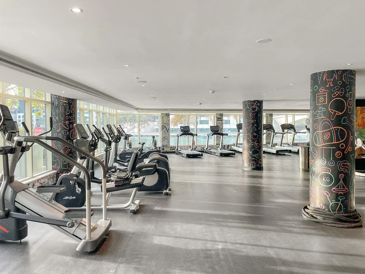 Un gimnasio amplio con equipos cardiovasculares junto a grandes ventanas.