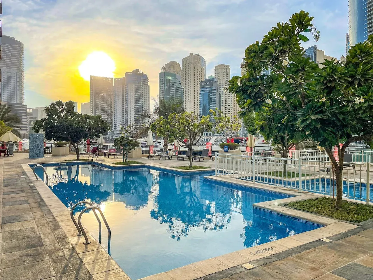 Una piscina al aire libre refleja el atardecer, con árboles y edificios altos cerca.