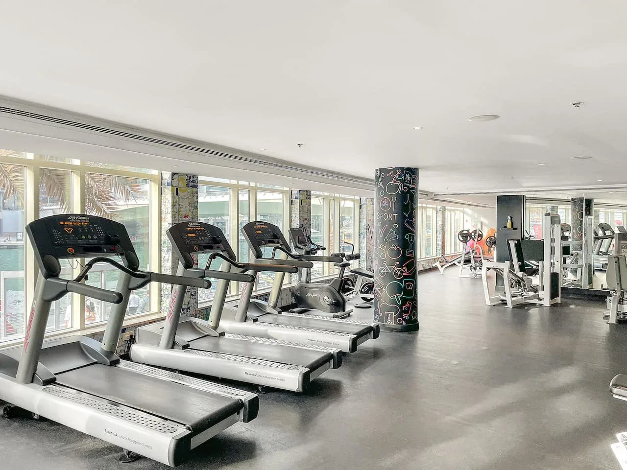 Un gimnasio con cintas de correr junto a grandes ventanas y equipo de ejercicio.