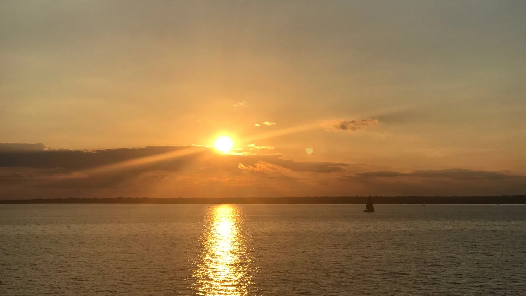 Sunset over calm water, with a small sailboat near the horizon.