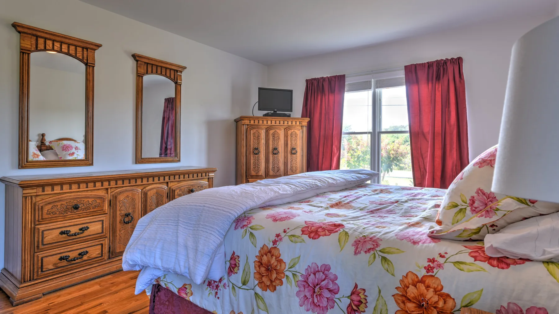 A bedroom with a floral bedspread, wooden dresser, and red curtains by the window.