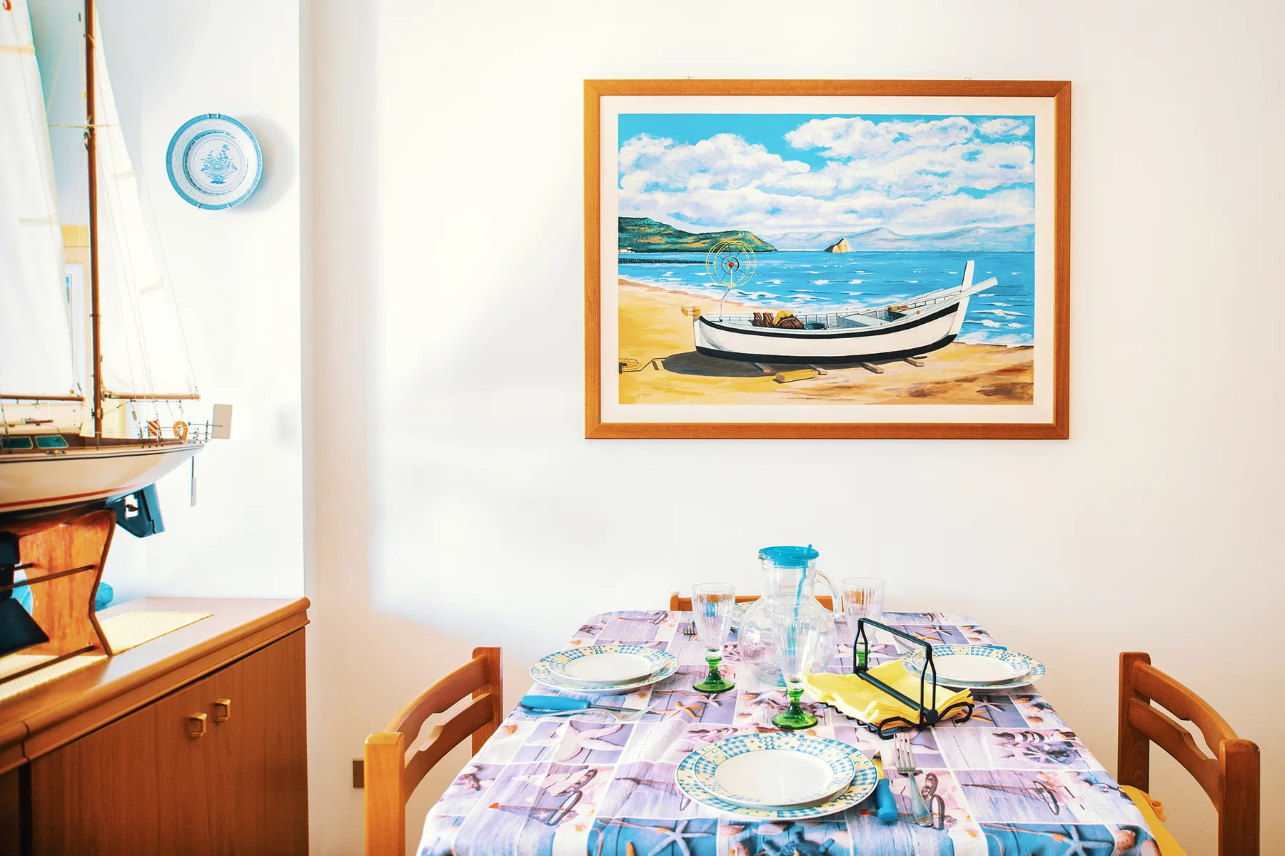 Un comedor con la mesa puesta y un cuadro enmarcado de un barco junto al mar.