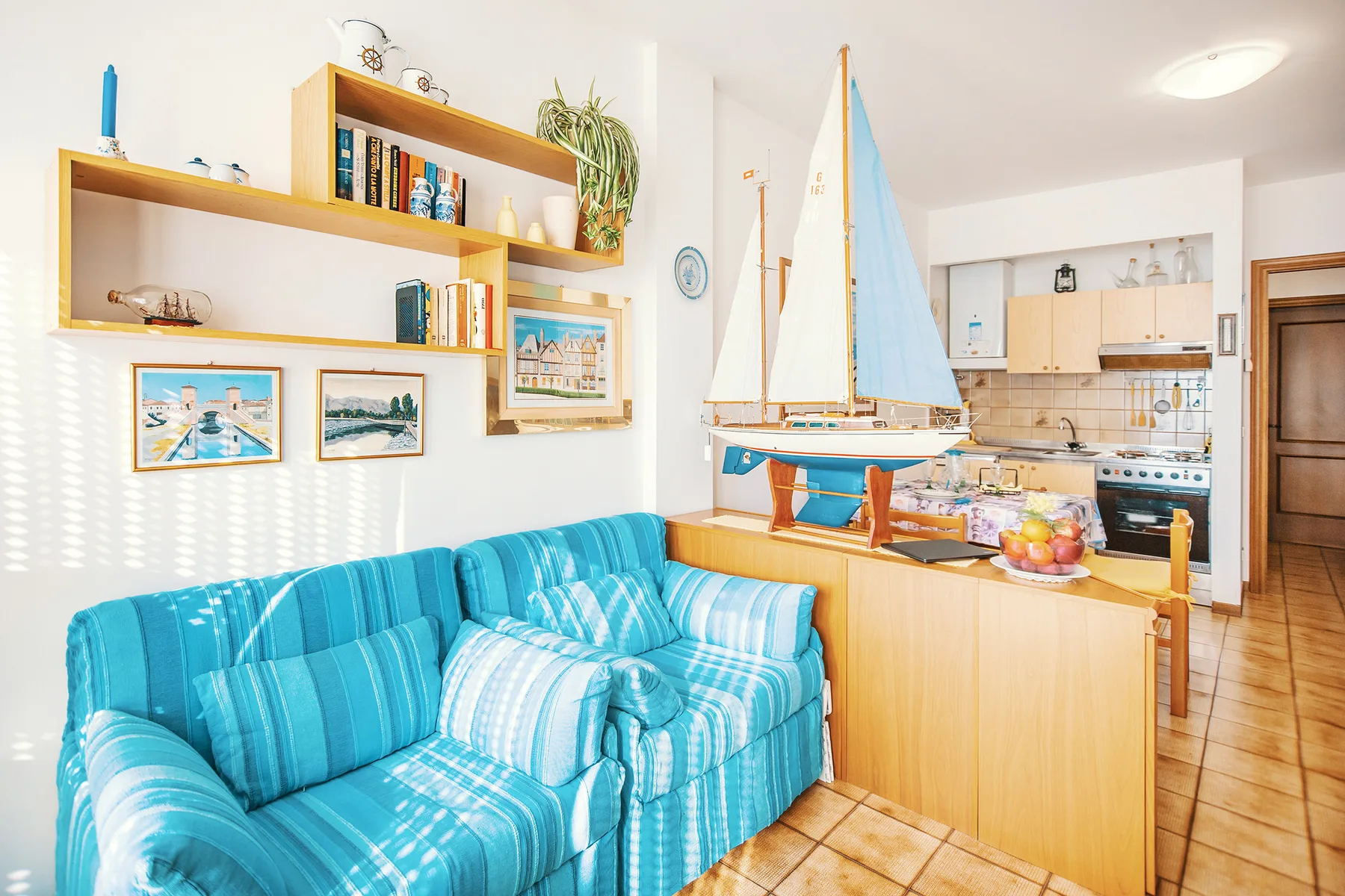 A bright living room with turquoise seating, a model sailboat, and an open kitchen.