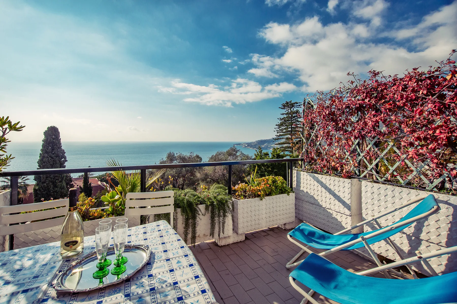 A sea-view terrace with outdoor seating, a table, and red-leaved climbing plants.