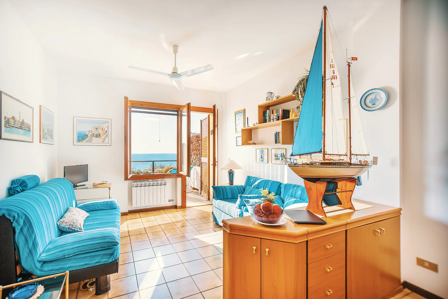 Una sala de estar luminosa con asientos turquesa, un mueble de madera y un velero a escala.