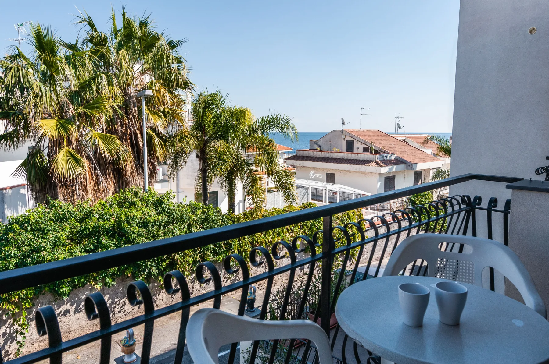 A balcony seating area overlooks palm trees, nearby rooftops, and the sea.
