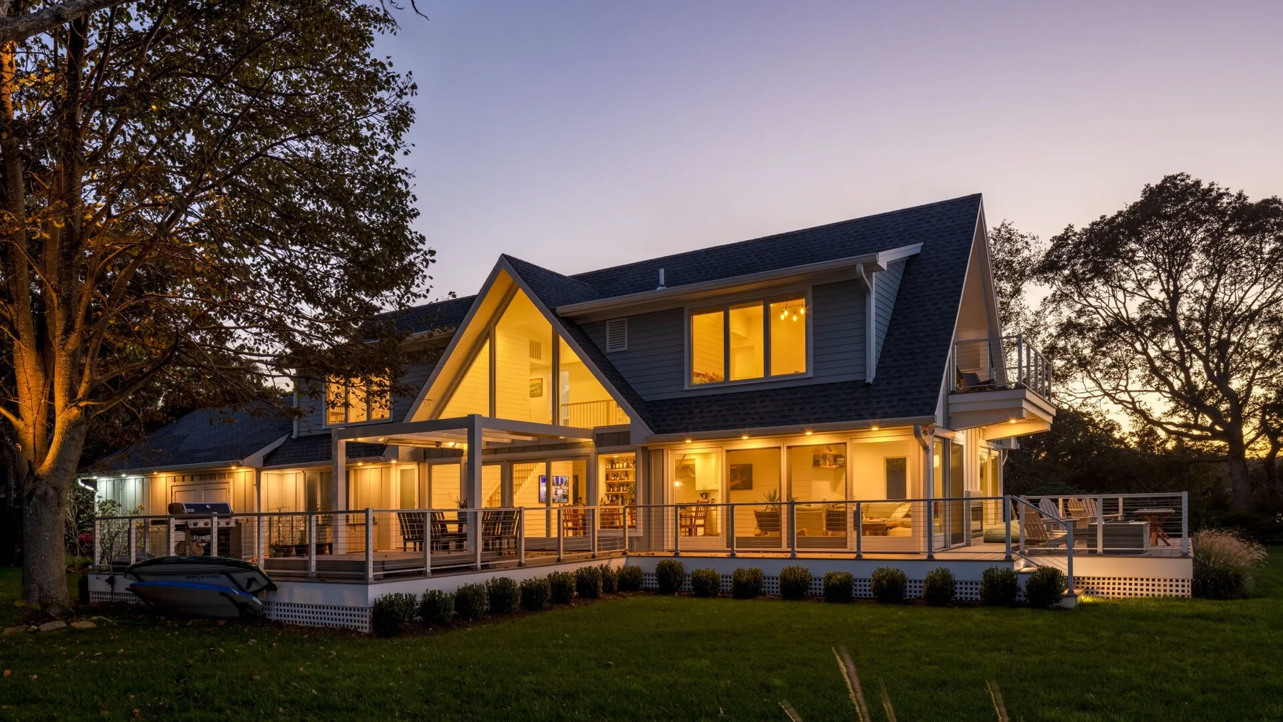 Una casa grande brilla con luces cálidas al anochecer, rodeada de árboles y césped.