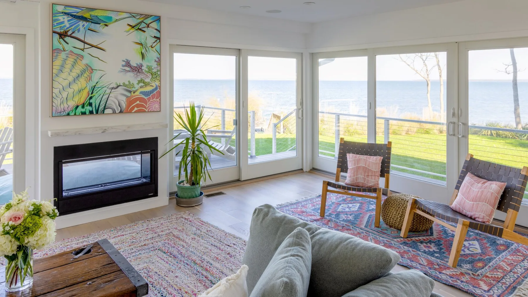 Una sala luminosa tiene una chimenea, asientos y grandes ventanas con vista al agua.
