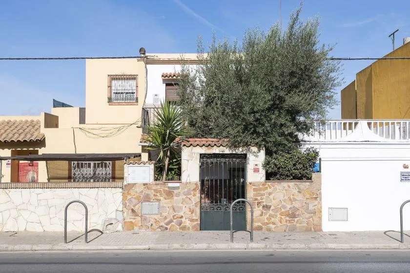 A residential building stands behind a stone wall and metal gate, with trees in front.