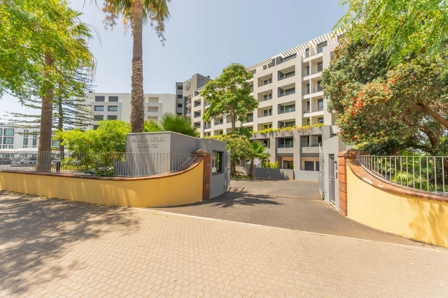 Un edificio de apartamentos se alza tras una entrada pavimentada rodeada de árboles.