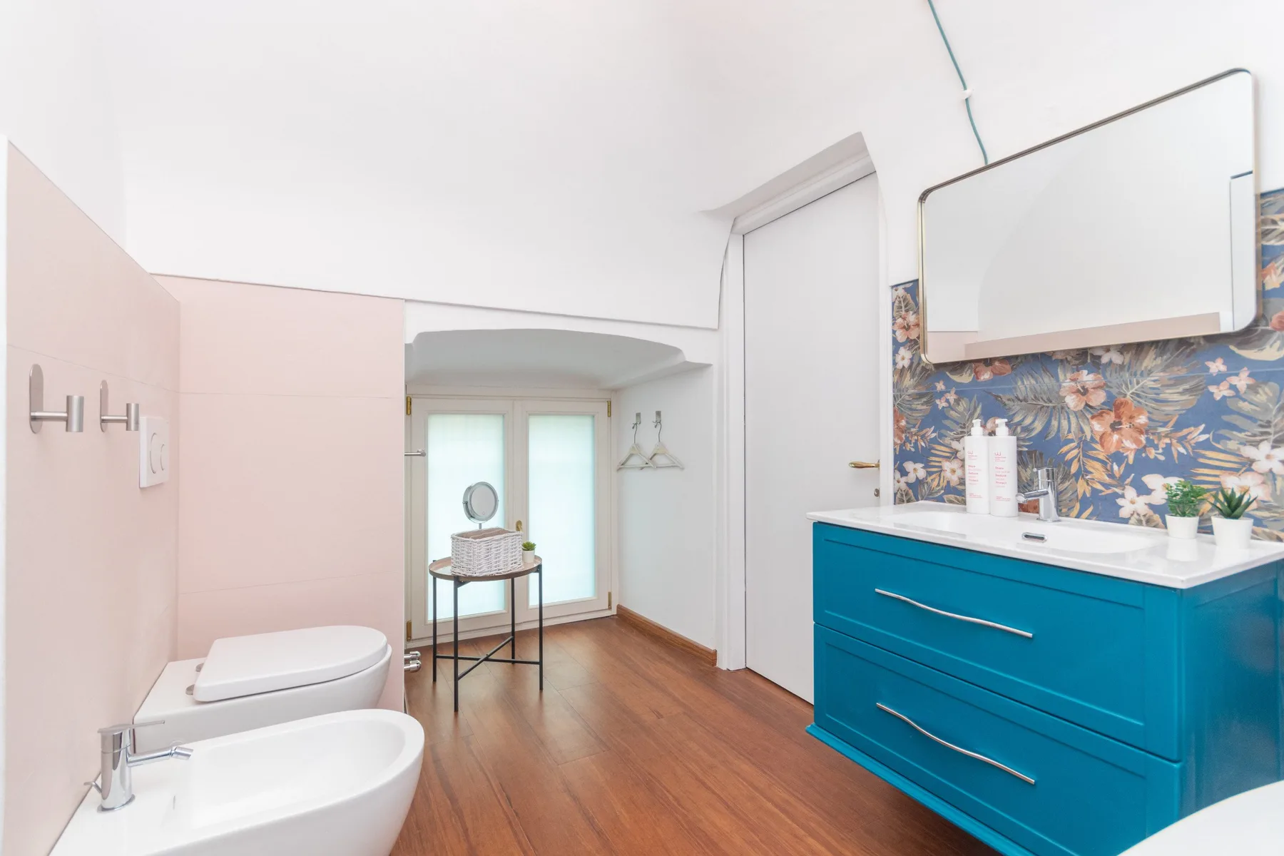 A bright bathroom with a turquoise vanity, floral wall covering, and wood flooring.