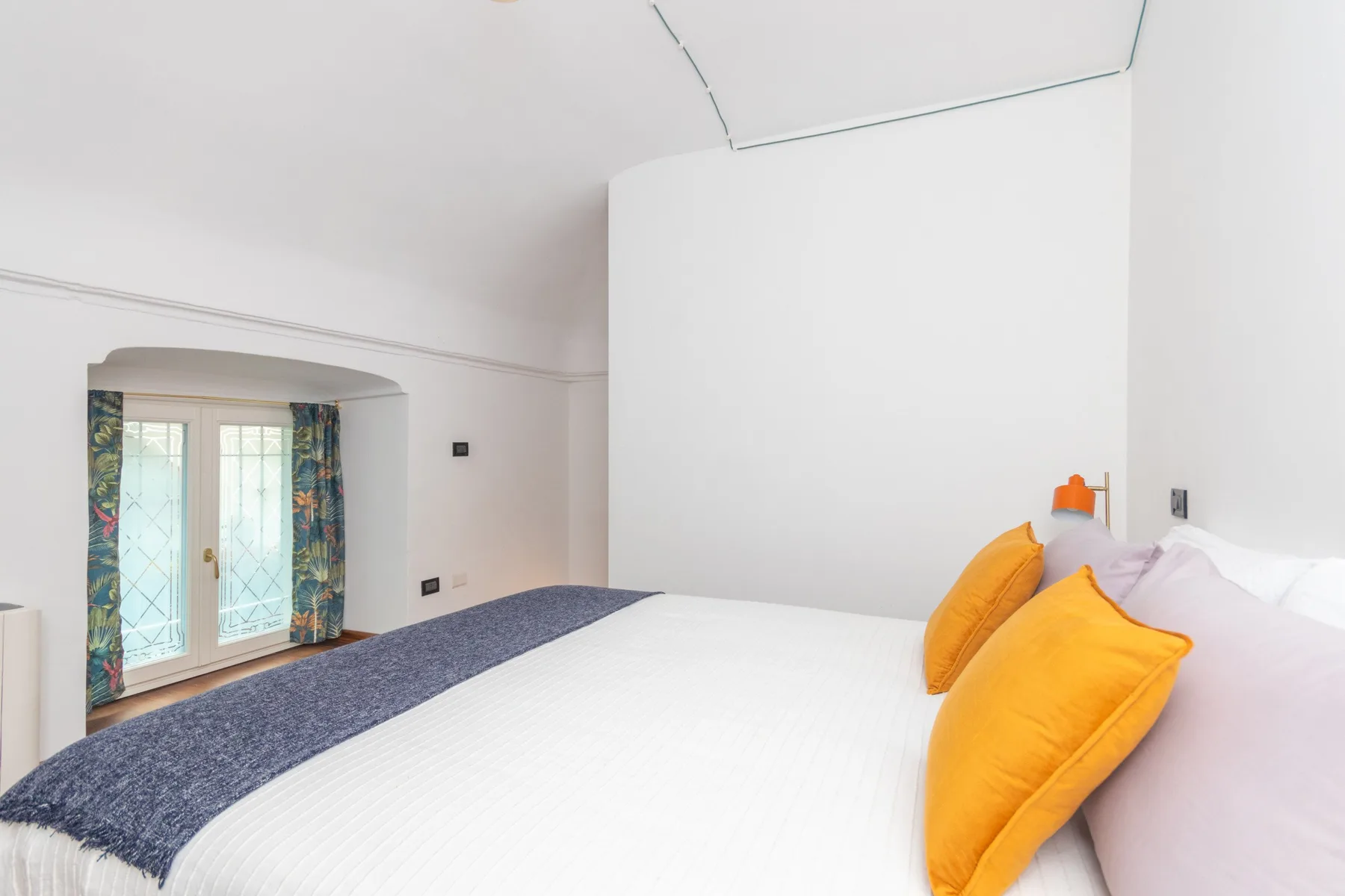 Un dormitorio con una cama blanca y almohadas naranjas junto a una ventana con cortinas estampadas.