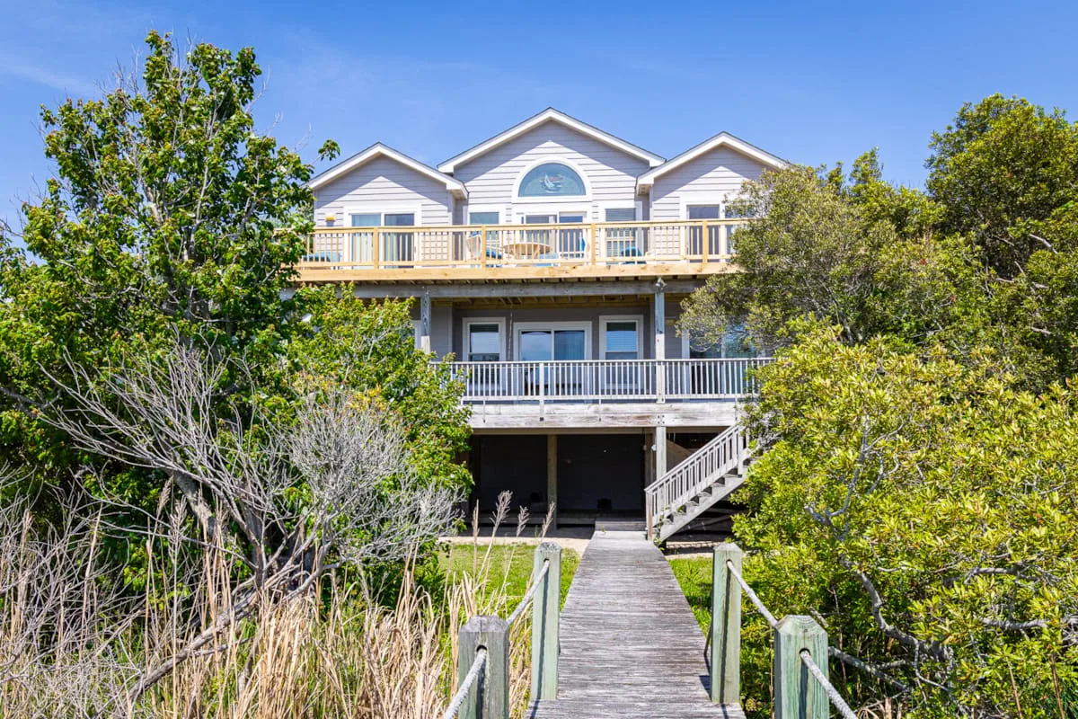A large coastal-style house with balconies stands among leafy trees.