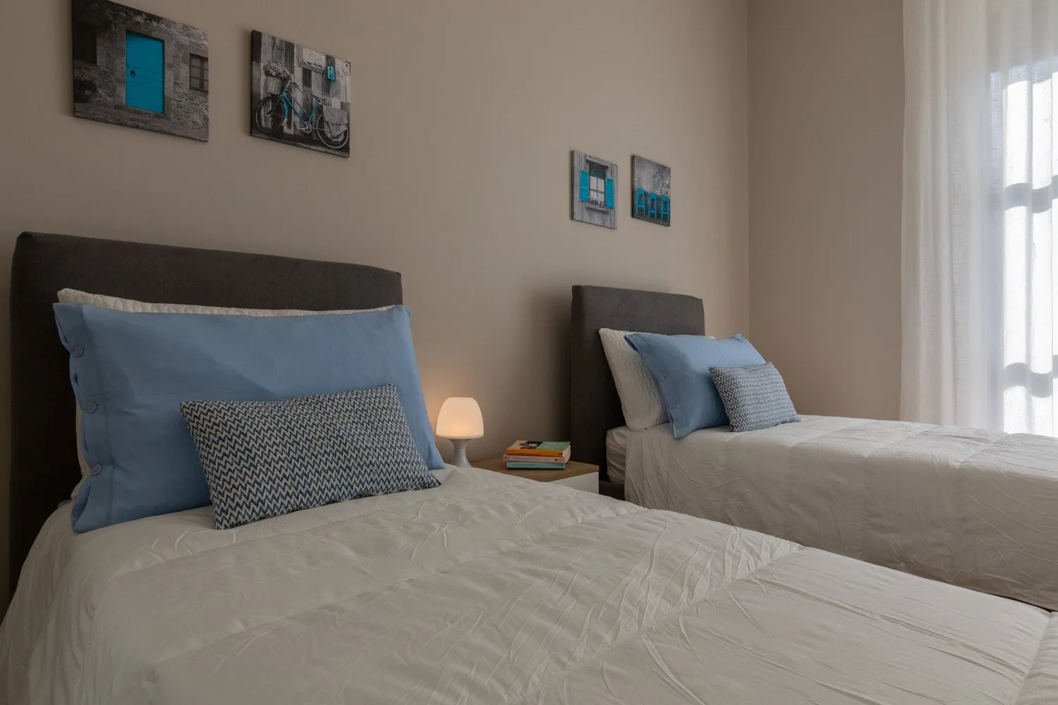 A bedroom with two beds, blue pillows, and framed artwork on the wall.