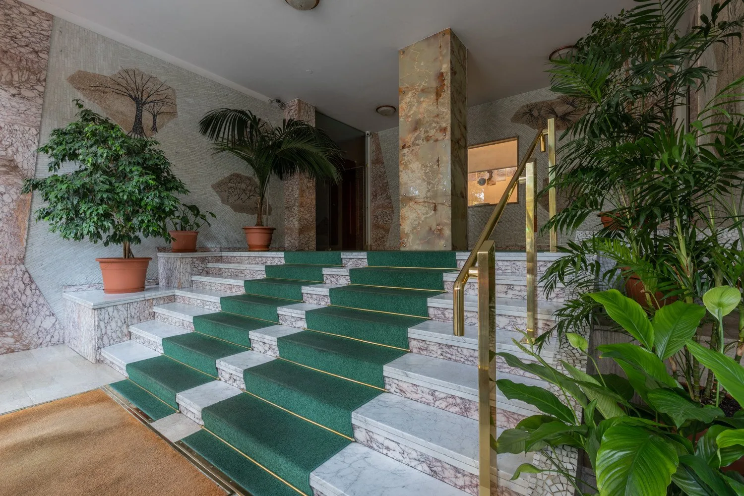 A carpeted staircase with green runners rises between marble walls and potted plants.