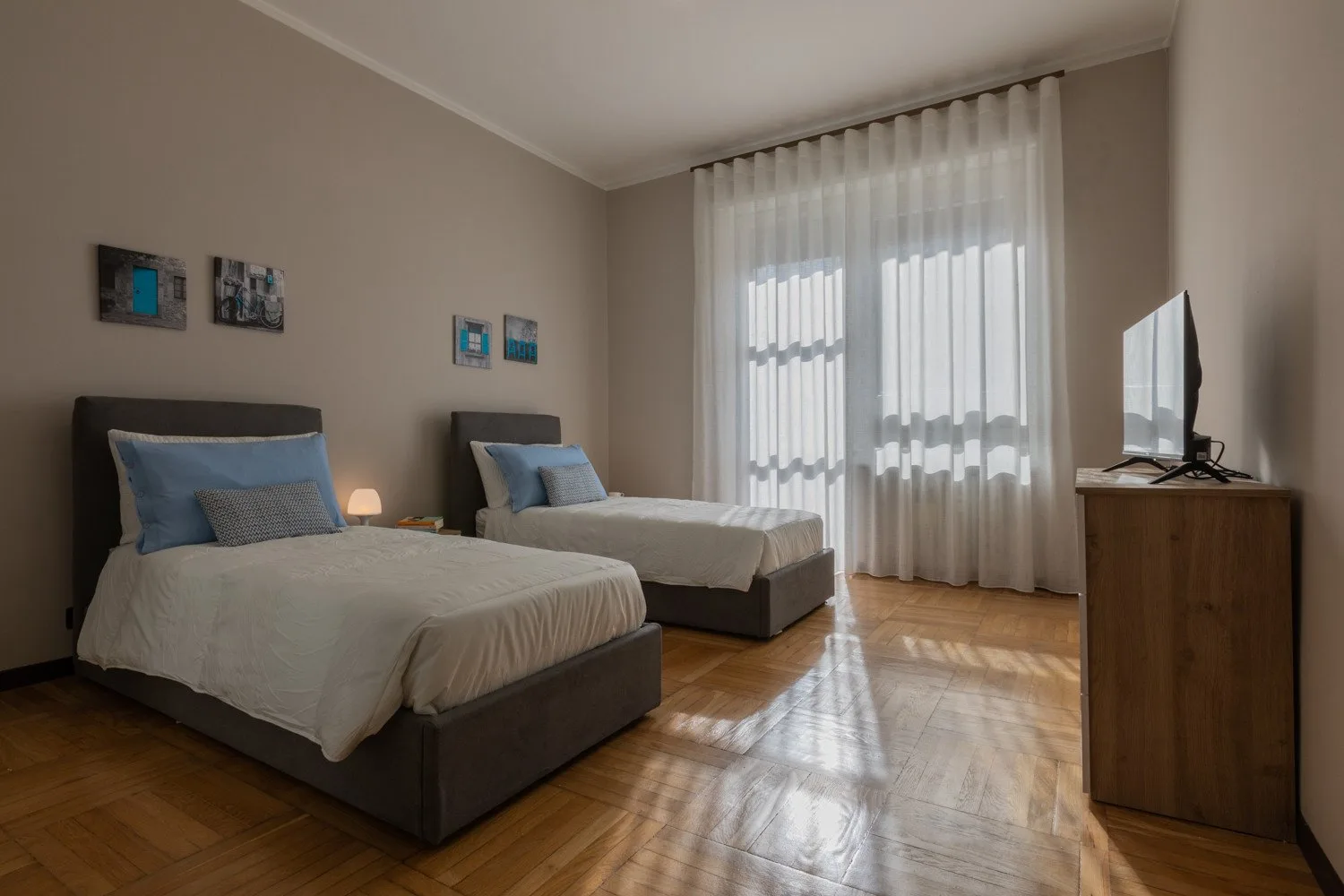 A bedroom with two beds, sheer curtains, and sunlight on the wood floor.