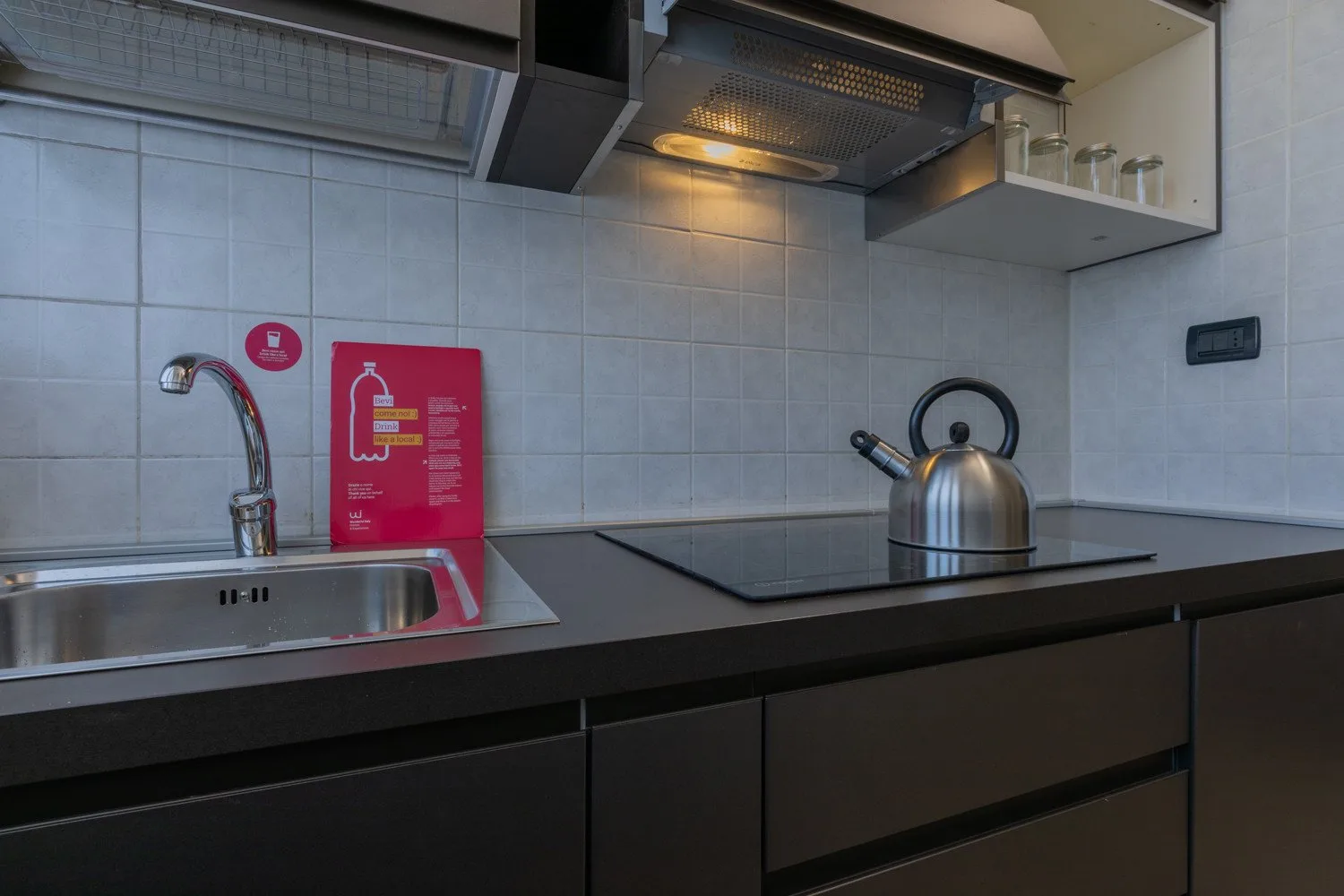 A kitchen counter with a sink, cooktop, and kettle beneath a range hood.
