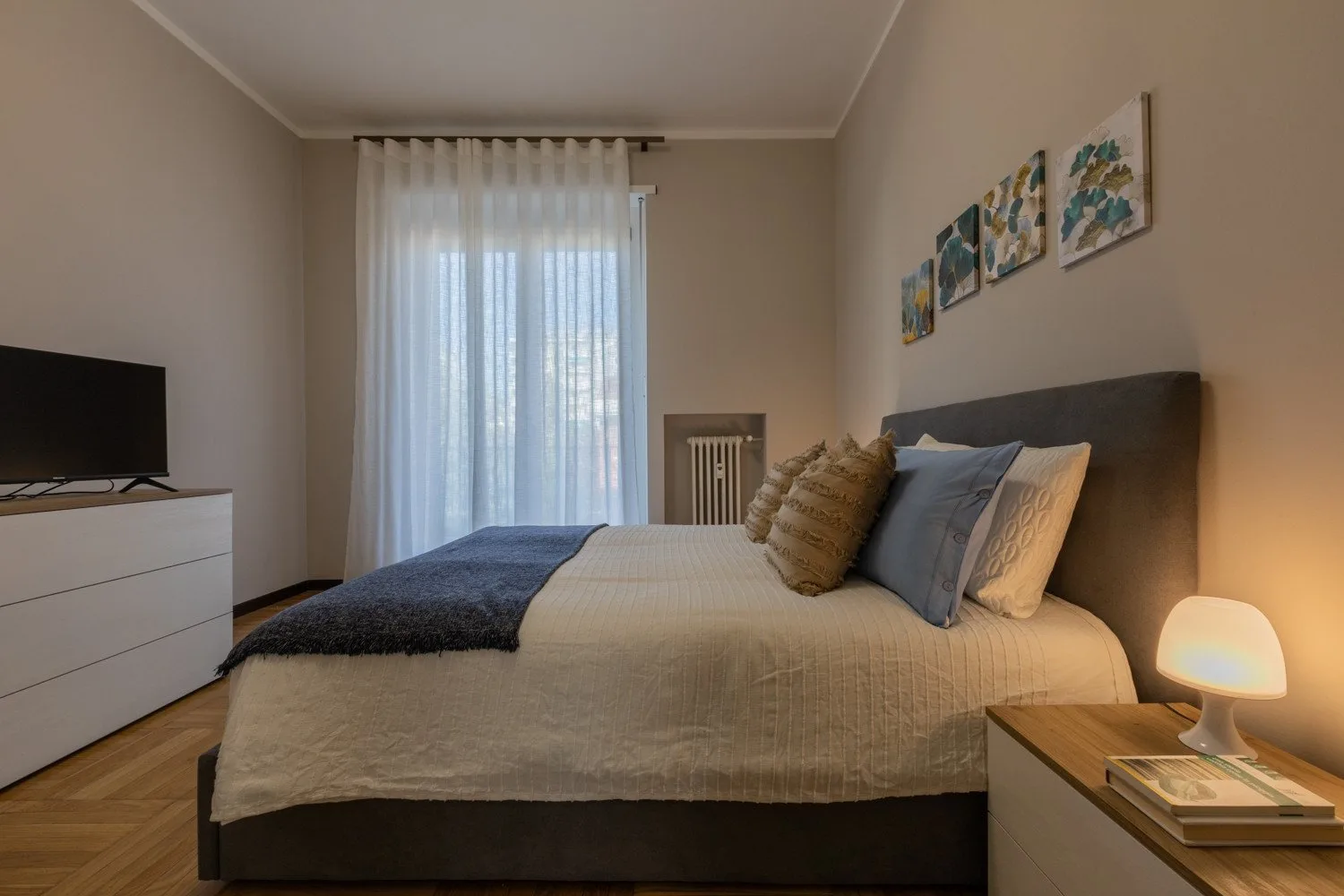 A bedroom with a neatly made bed, soft curtains, and framed artwork.