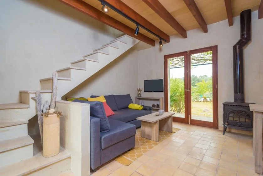 Una sala de estar con sofá seccional, escalera y puertas de vidrio frente a la vegetación.