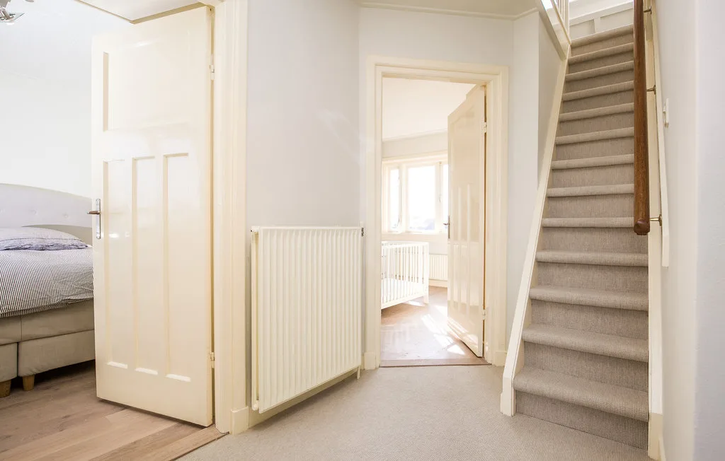 Un pasillo luminoso con una escalera, un radiador y una puerta abierta a un dormitorio.
