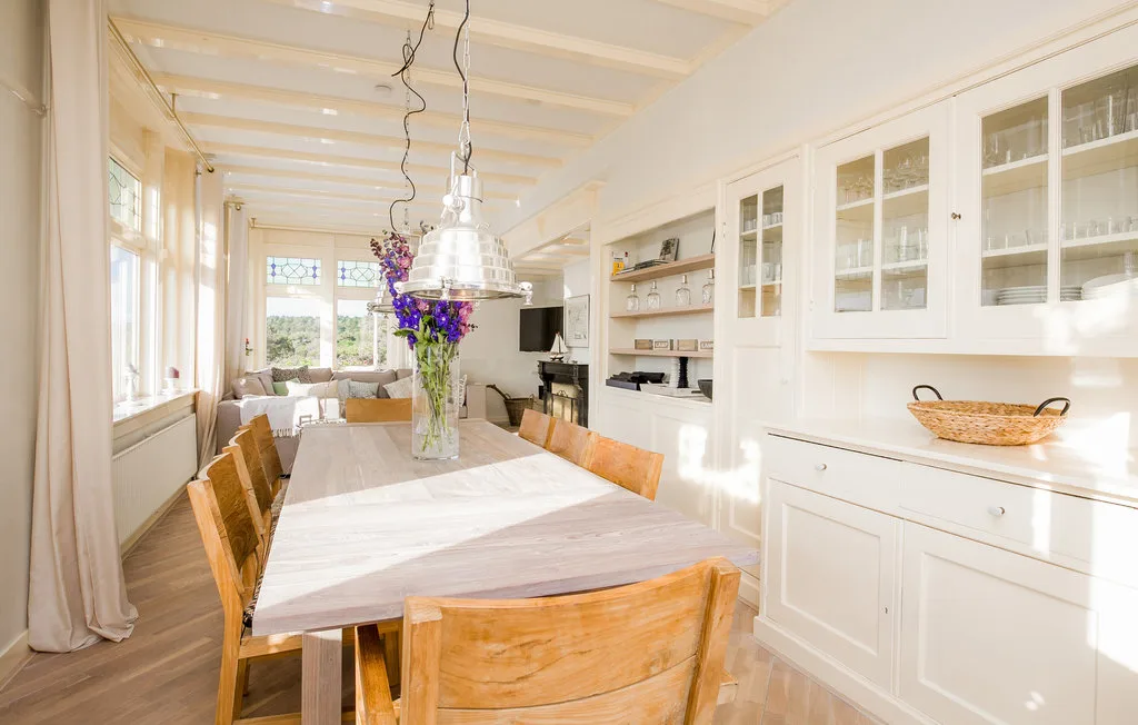 Un comedor luminoso con una mesa larga de madera y flores bajo una lámpara colgante.