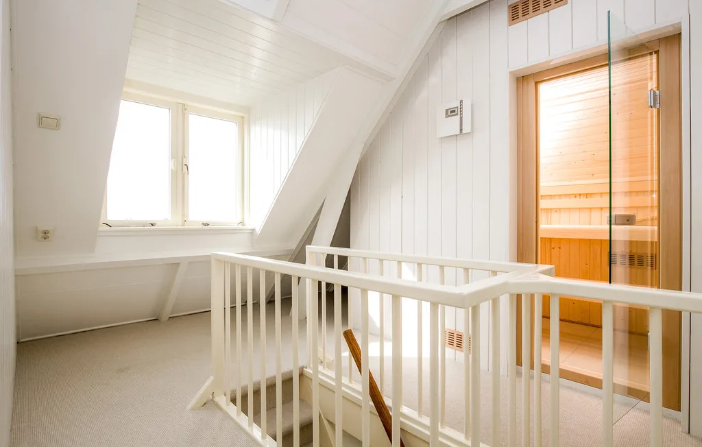 Un descansillo abuhardillado luminoso con barandilla blanca y una entrada de madera a la sauna.