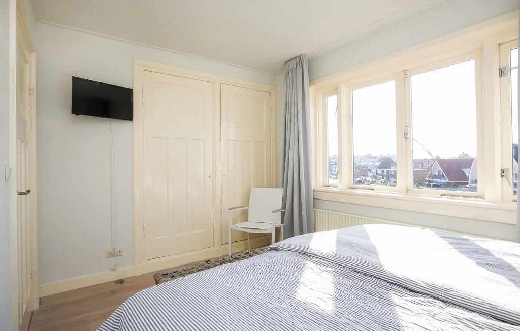 Un dormitorio luminoso con una cama junto a grandes ventanas y un televisor en la pared.