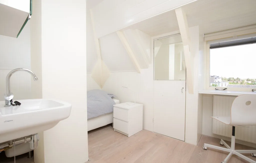 Una habitación luminosa con cama, escritorio y lavabo bajo un techo inclinado.