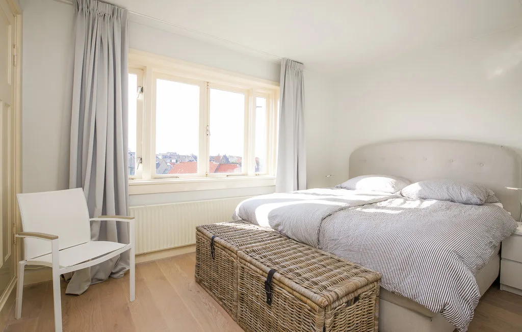 Un dormitorio luminoso con una ventana grande, cortinas y un banco de mimbre junto a la cama.