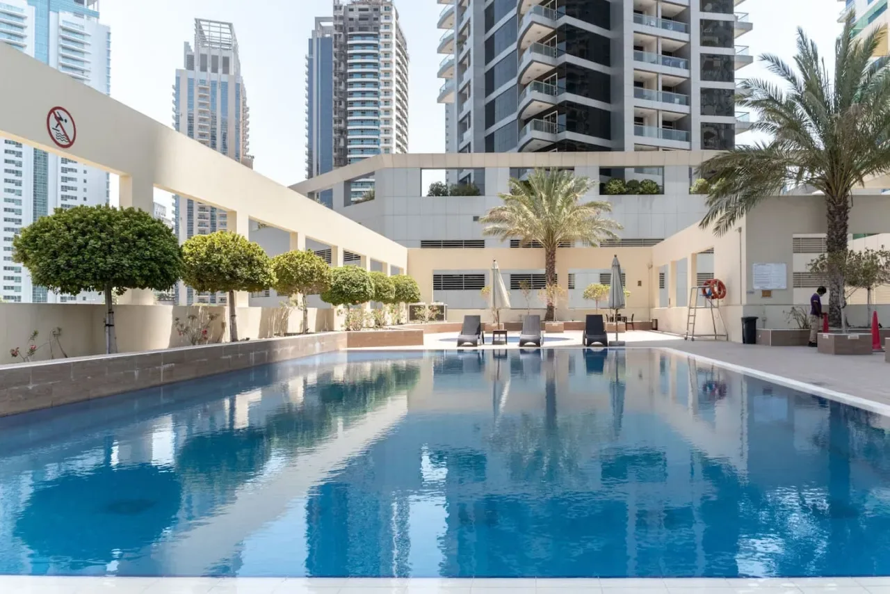 Una piscina está junto a una zona ajardinada con asientos y edificios modernos de gran altura.