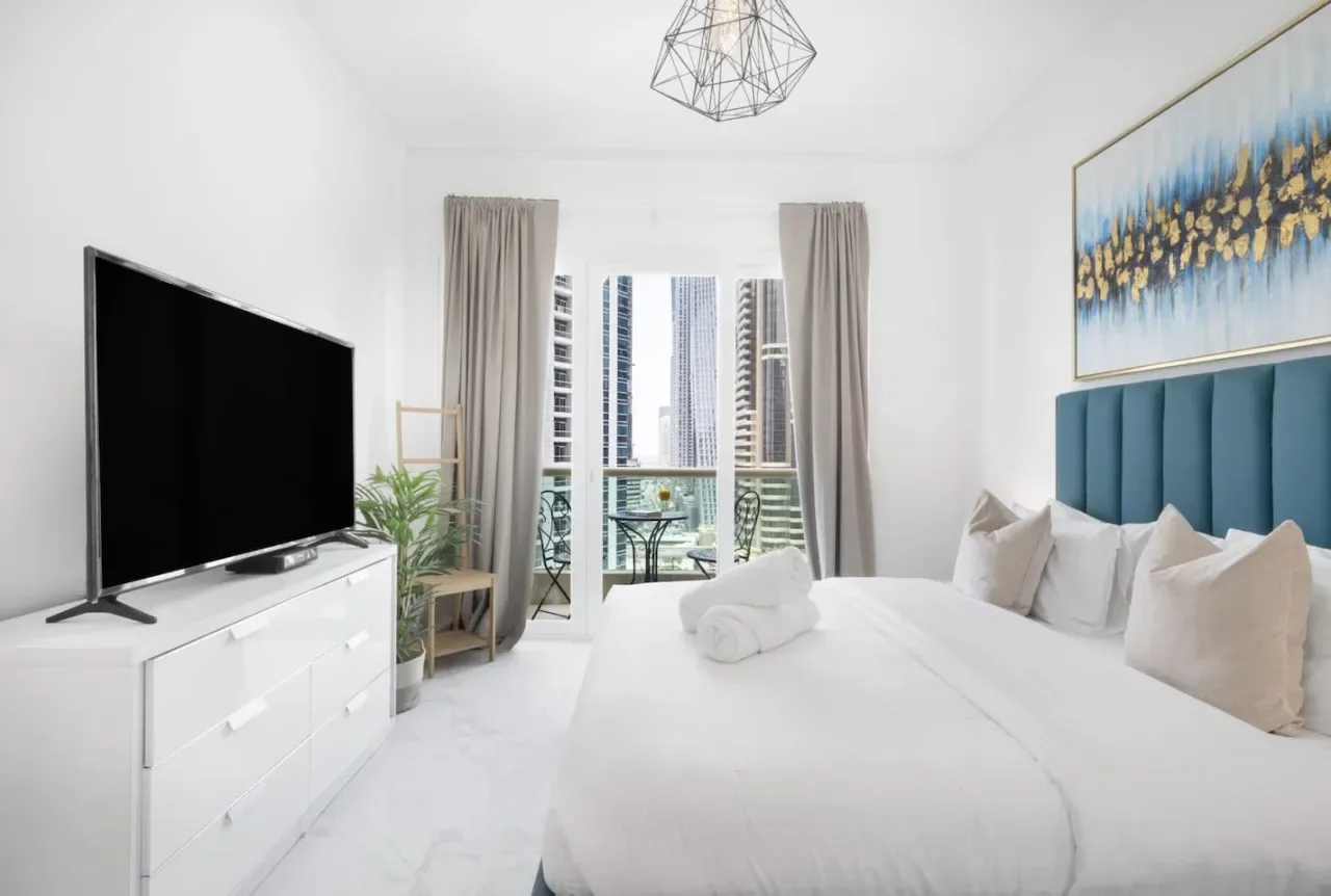 Un dormitorio luminoso con una cama grande, un televisor y ventanas con vistas a edificios cercanos.