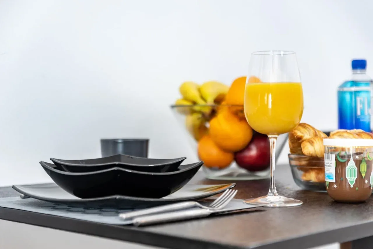 Una mesa está preparada con platos, cubiertos, fruta y un vaso de zumo de naranja.