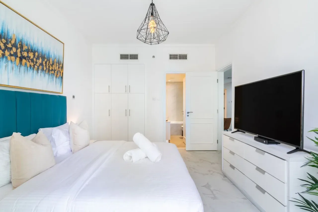 Un dormitorio luminoso con una cama blanca, cabecero verde azulado y televisor.