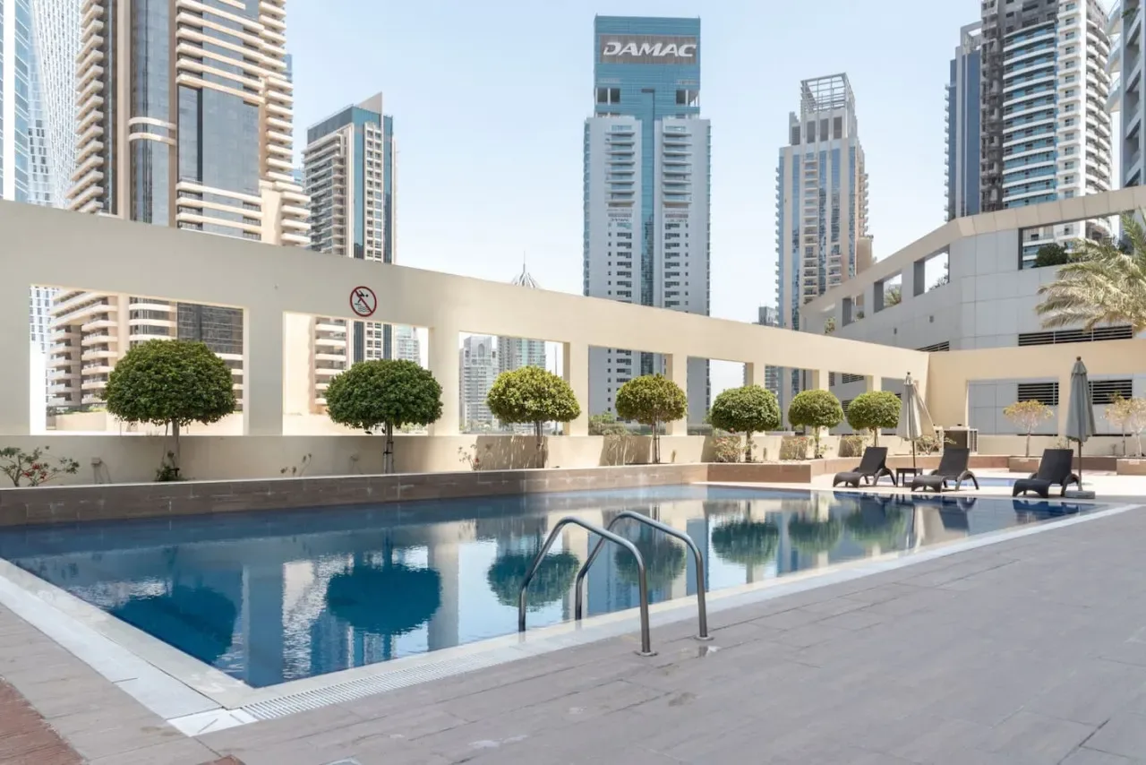 Una piscina al aire libre está junto a una terraza pavimentada, con edificios altos al fondo.