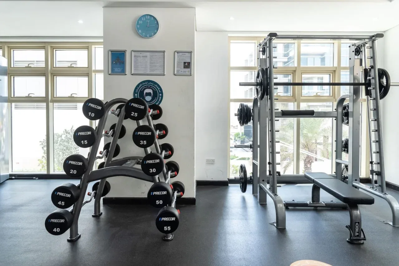 Un gimnasio luminoso con un soporte para pesas y equipo de entrenamiento junto a grandes ventanas.