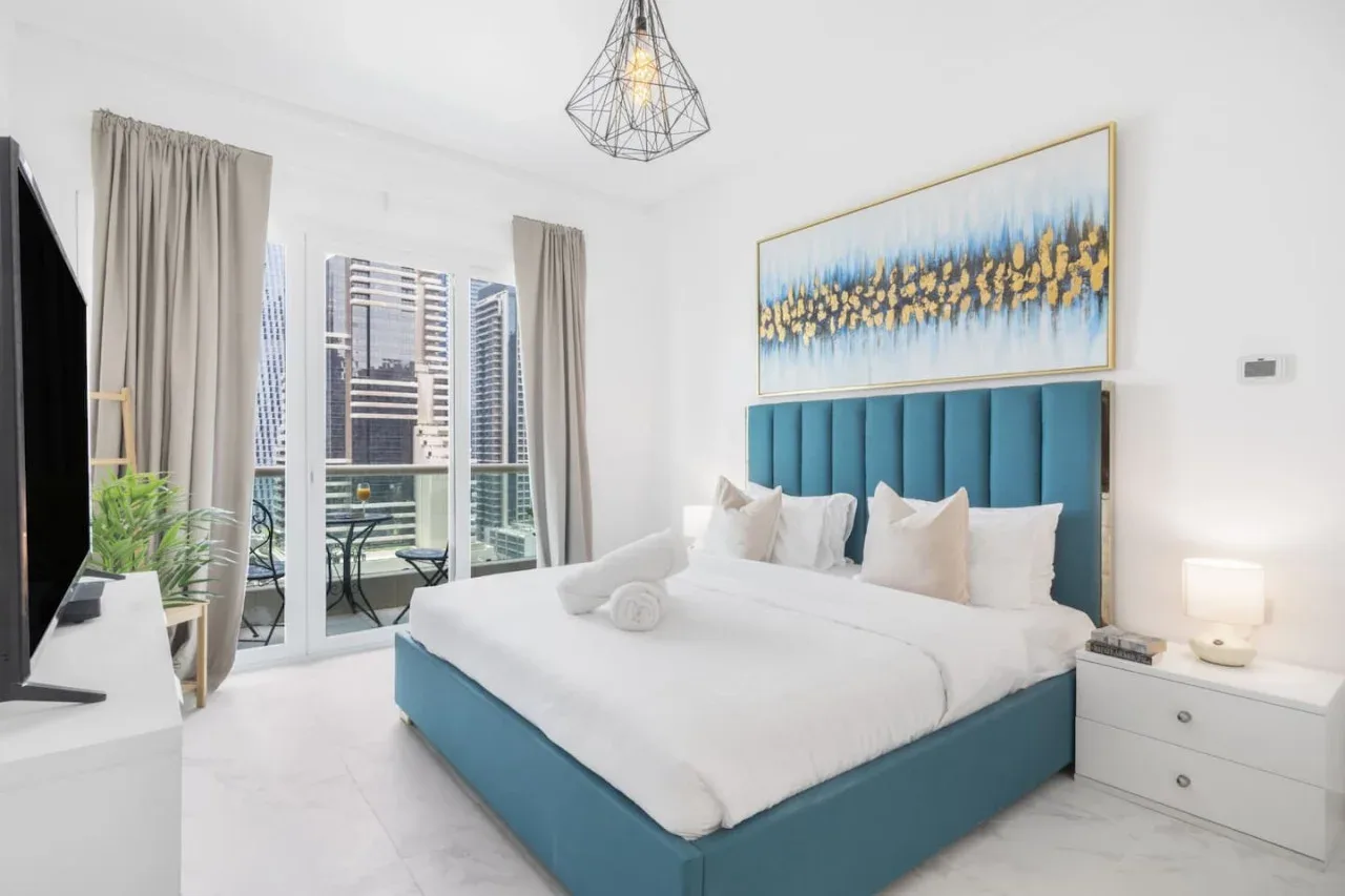 Un dormitorio luminoso con una cama grande, cabecero verde azulado y ventanas con vistas a edificios cercanos.