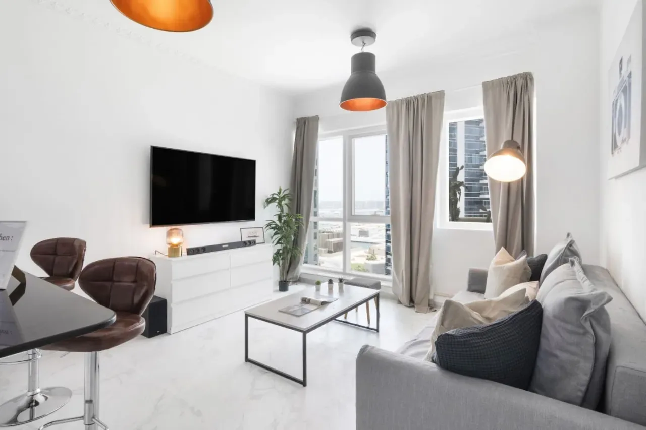 Una sala luminosa con un sofá gris, televisor y ventanales.