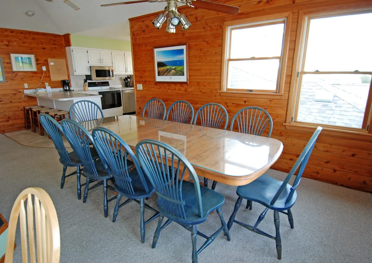 A dining area with a long table and blue chairs beside large windows.