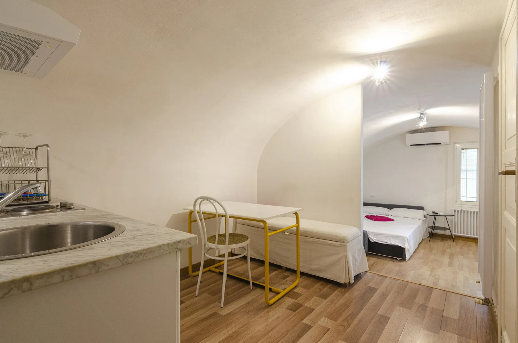 Una pequeña cocina se abre a un dormitorio con una cama y una mesa pequeña.