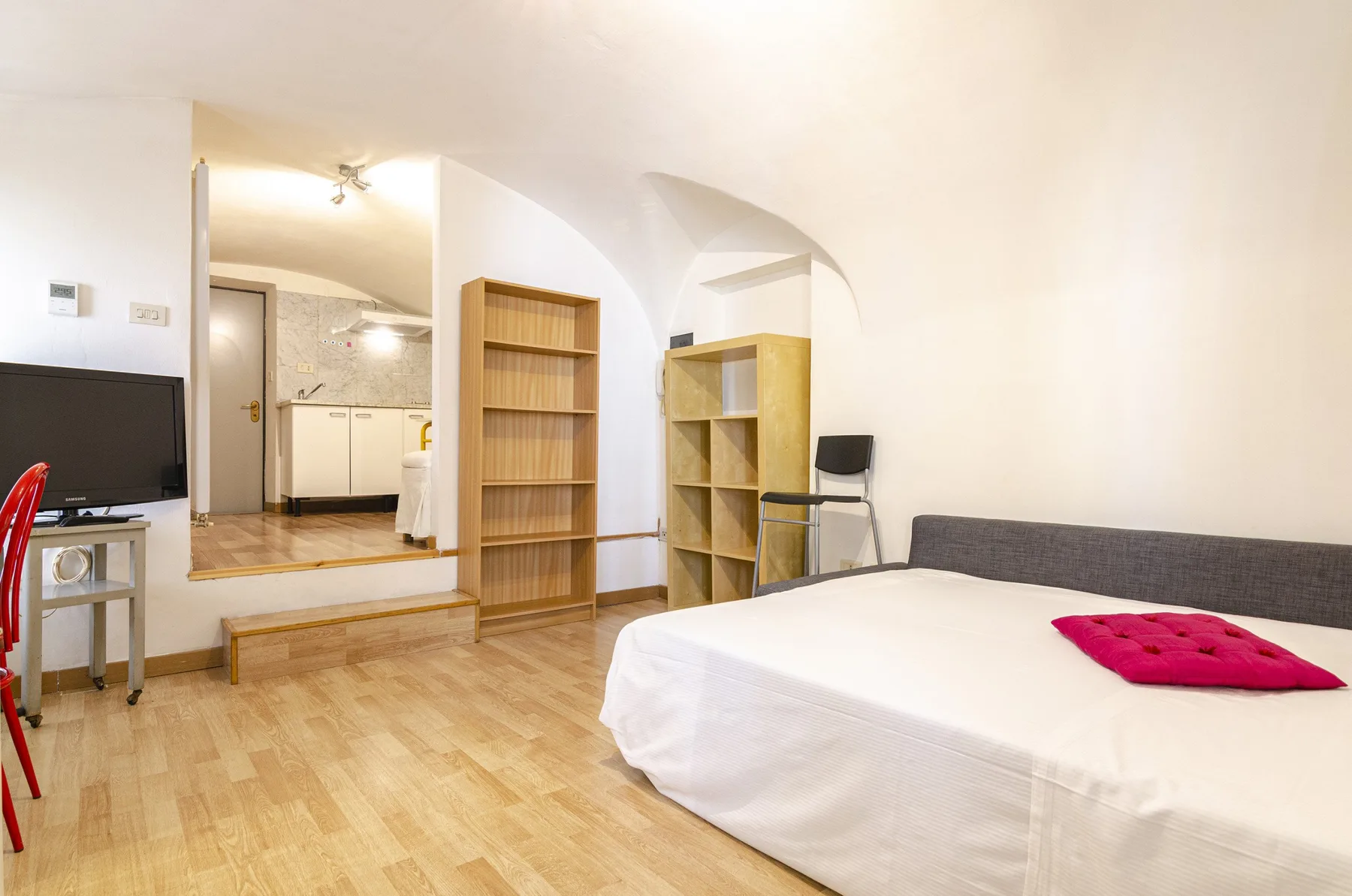 Un dormitorio con una cama grande, estanterías abiertas y una pequeña zona de cocina al fondo.