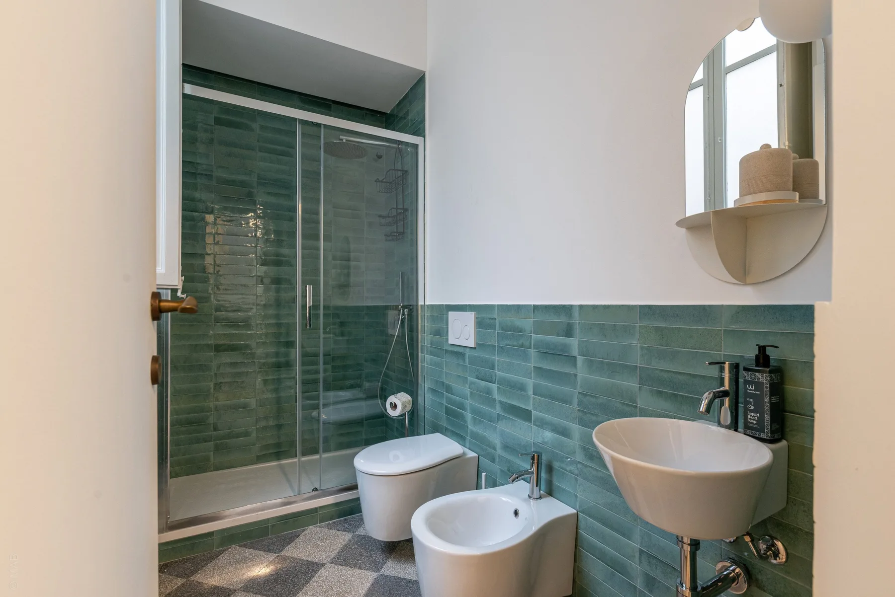 A bathroom with green wall tiles, a glass shower, and a wall-mounted sink.