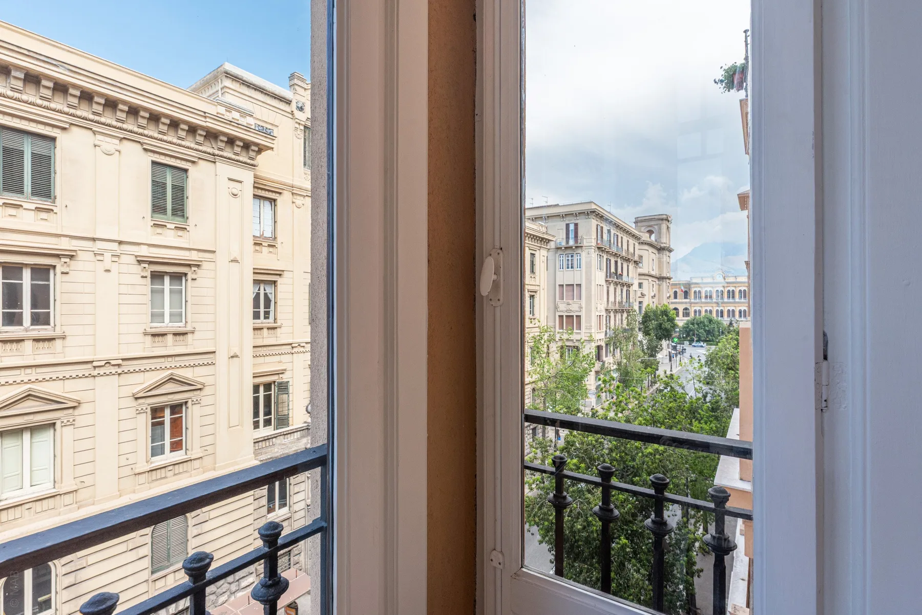 A view through an open window overlooks leafy trees and pale city buildings.