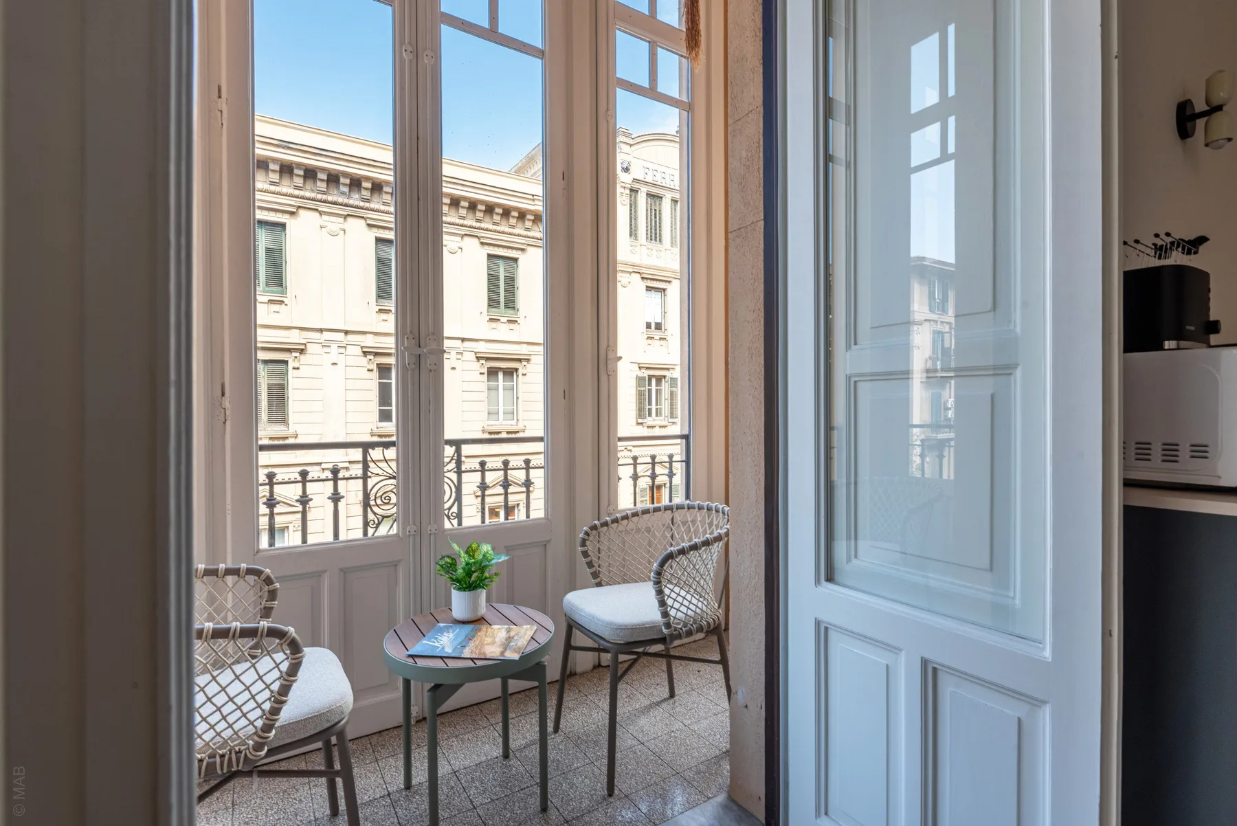 A bright balcony seating area overlooks a neighboring building through tall windows.