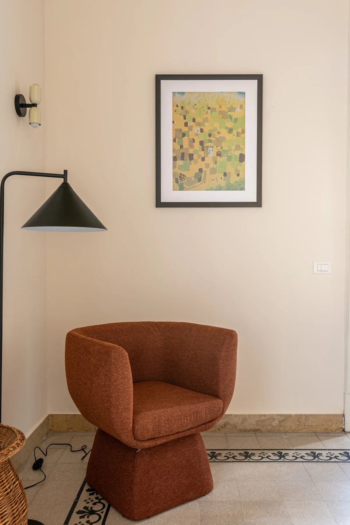 A rust-colored upholstered chair sits beneath framed artwork beside a wall-mounted lamp.