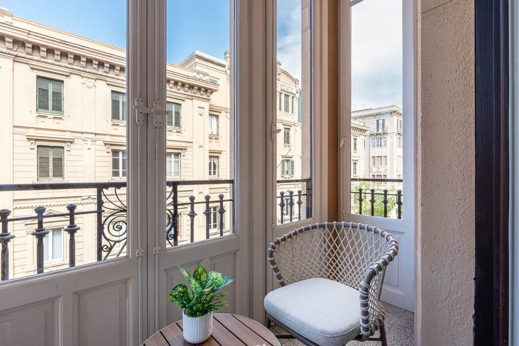 A bright enclosed balcony has tall windows, a small table, and a cushioned chair.