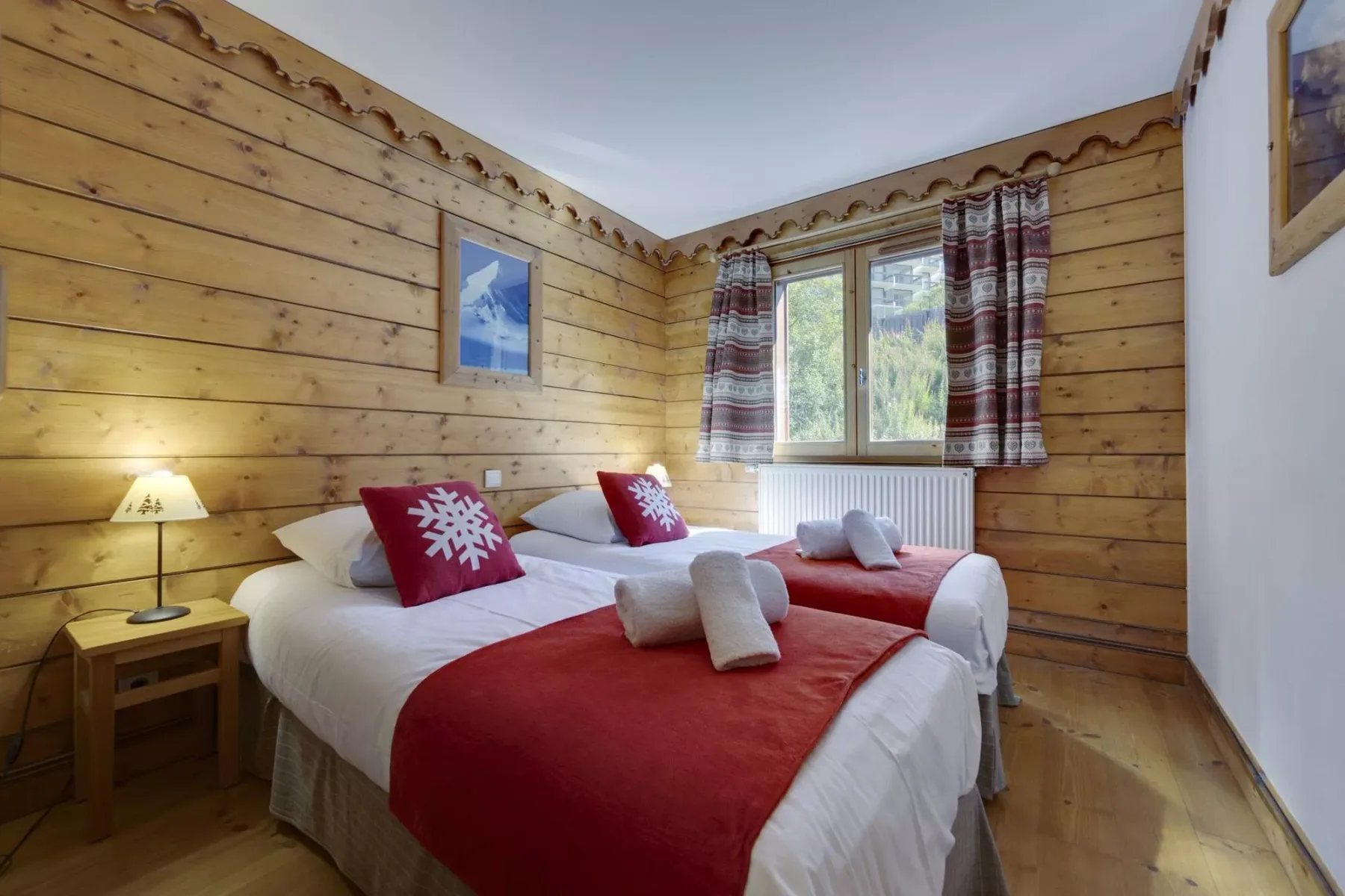 Dormitorio con paredes de madera y camas vestidas con ropa blanca y roja junto a una ventana.